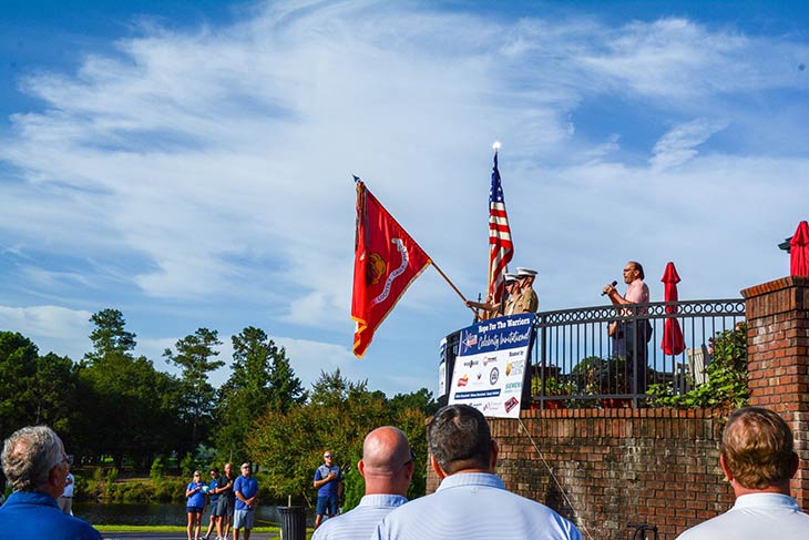 Photo of a man speaking into a microphone at an Hope For The Warriors 2021 with Marines holding flags